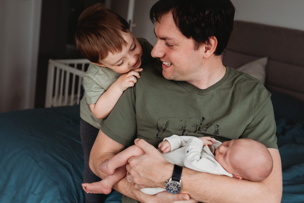 otec držící novorozeného syna pozoruje staršího, který ho objímá
