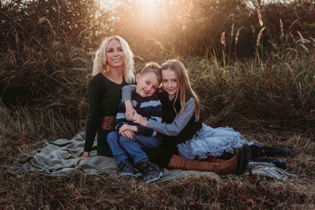 Maminka sedí na dece se svou dcerou a se synem mezi vysokou suchou trávou.