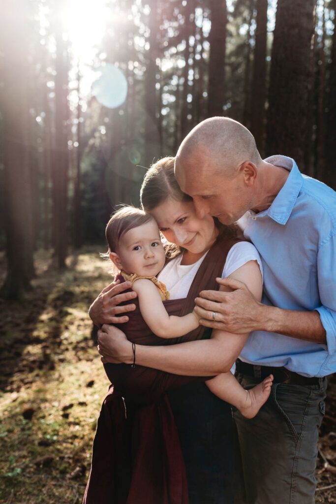 Rodina s dcerou v šátku stojí mezi stromy v lese a prosvítá na ně slunce.