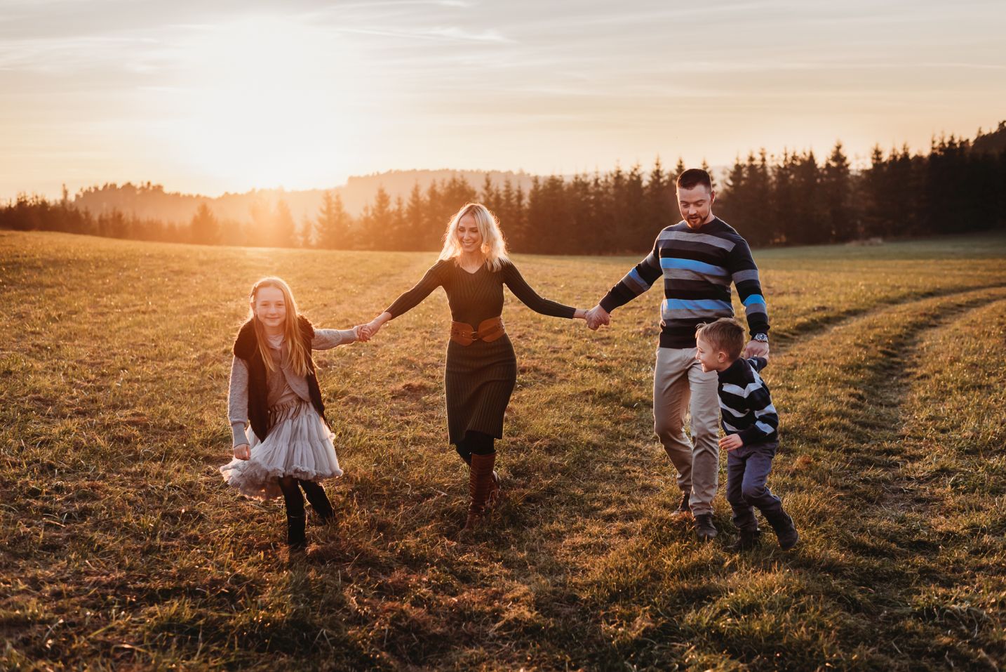 Rodina se drží za ruce a kráčí při západu slunce po louce.
