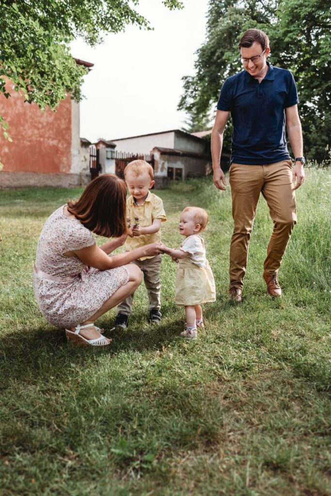 Starší syn přinesl mamince květinu a pozoruje to mladší dcera i tatínek.
