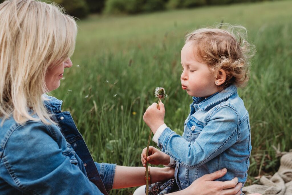 Holčička sedí na matčiných nohách a fouká do pampelišky.